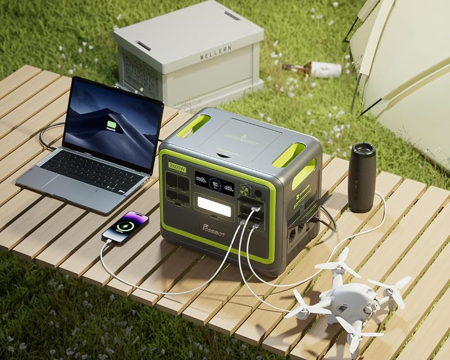 A portable power station with gray and lime green panels sits on a wooden slatted picnic table outdoors on grass, connected to a laptop, phone, speaker, and drone. A white storage box and part of a tent are visible in the background.